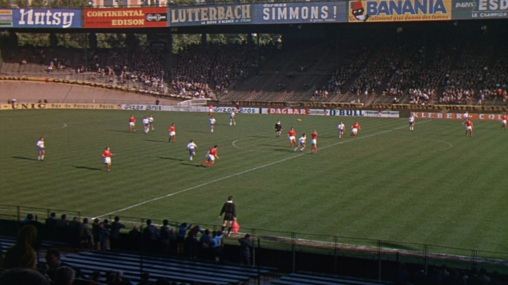 A soccer match in progress at a stadium, featuring players in red and blue jerseys on a green field, with spectators in the stands and advertisements in the background.