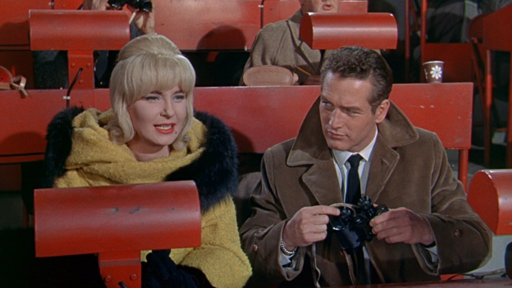 A blonde woman in a yellow coat and a man in a brown coat sit in a red stadium seating area, with the woman smiling and the man holding a camera.