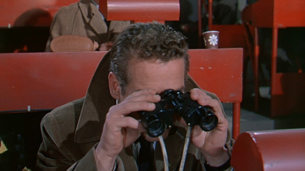 A man wearing a coat gazes through binoculars in a stadium filled with red seats.