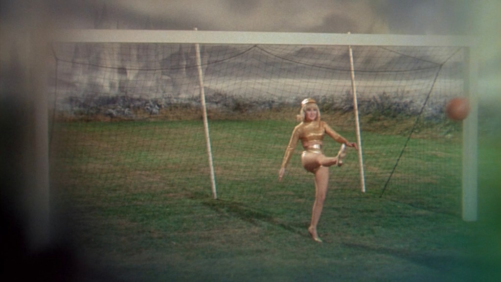 A woman in a shiny gold outfit plays soccer, lifting one leg to kick a ball towards a goalpost on a grassy field.
