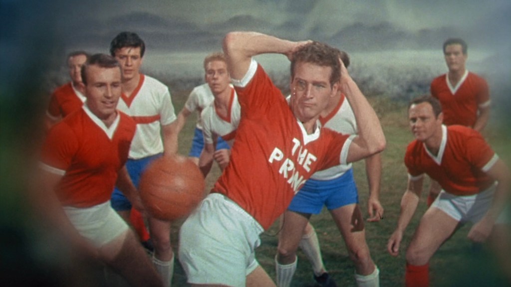 A group of male soccer players in red and white uniforms on a field, with one player in a red shirt throwing a basketball, while others watch attentively.