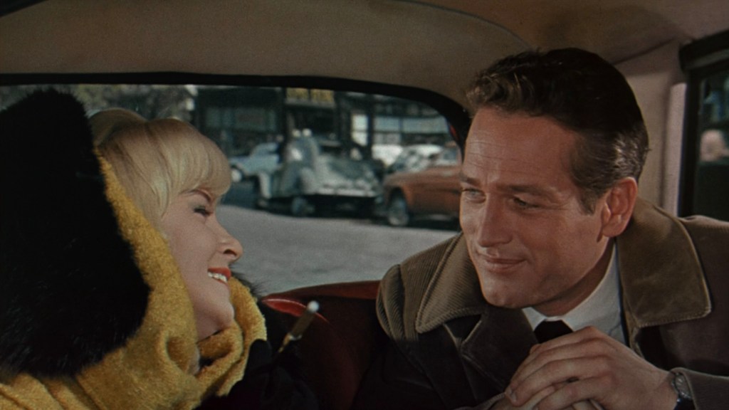 A man and a woman smiling at each other inside a car, with a city street visible in the background.