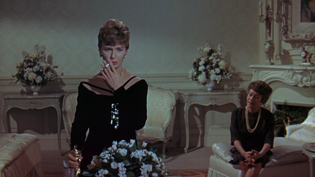 A glamorous woman in a black dress with a cigarette in hand stands thoughtfully in a luxurious room, while an older woman sits nearby, observing her with a concerned expression.