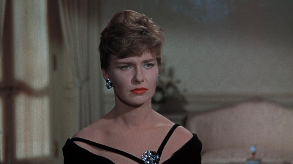 A woman in an elegant black dress with a low neckline, featuring a decorative brooch, stands with a serious expression. She has short, styled hair and bold red lipstick, set against a softly lit background.