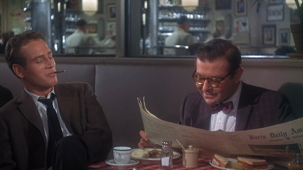 Two men seated at a table in a cafe, one with a cigarette and the other reading a newspaper. A cup and a small dish are on the table, along with a sandwich.