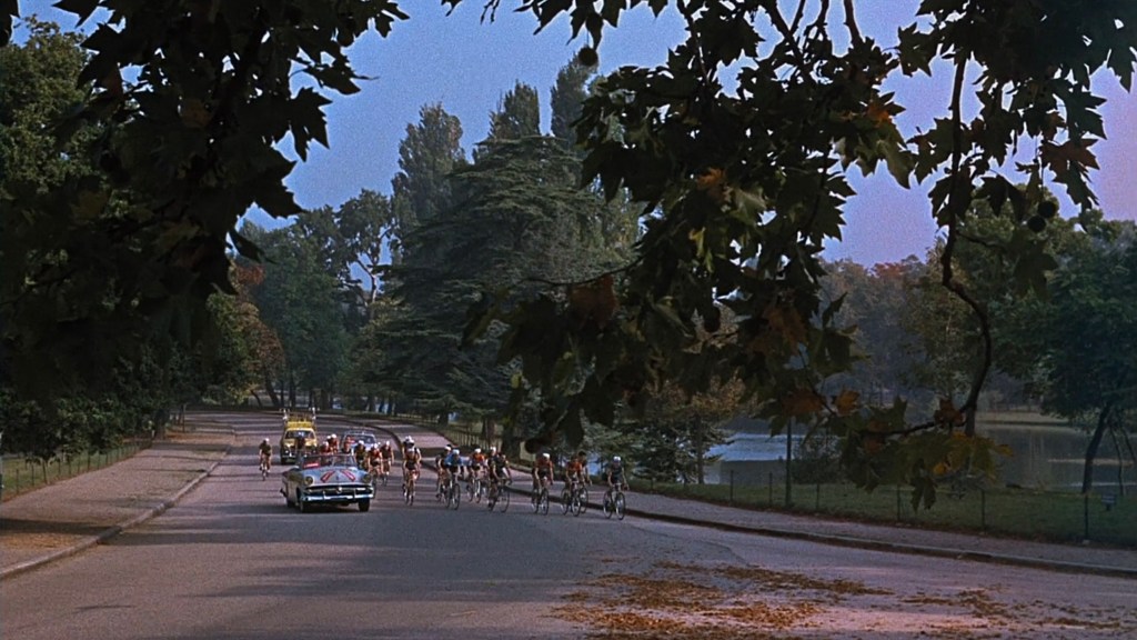 A group of cyclists riding along a tree-lined road, with a car following behind and a scenic view of a lake in the background.