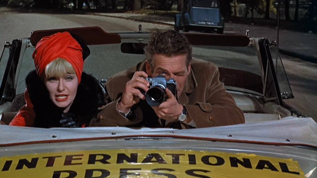 A woman with a red headscarf and black coat sits in a convertible car, looking surprised while a man beside her aims a camera at the scene outside. The car's dashboard displays a sign that reads 'INTERNATIONAL PRESS'.