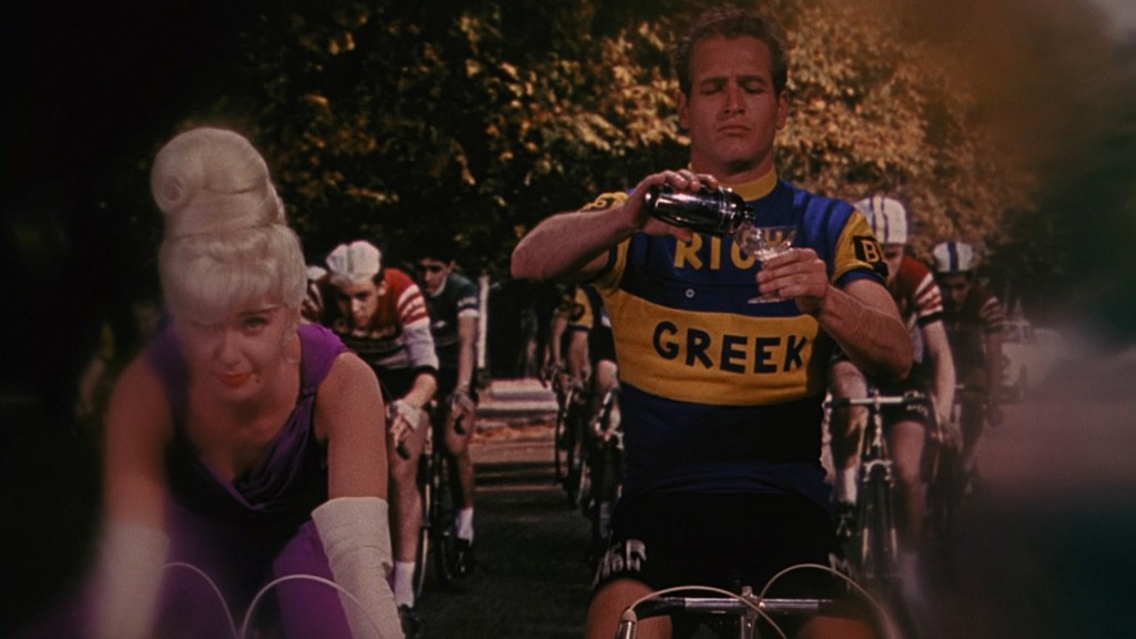A male cyclist in a blue and yellow jersey pours a drink from a bottle into a glass, while a female figure with a blonde updo in a purple outfit watches him. In the background, multiple cyclists are riding on a path.