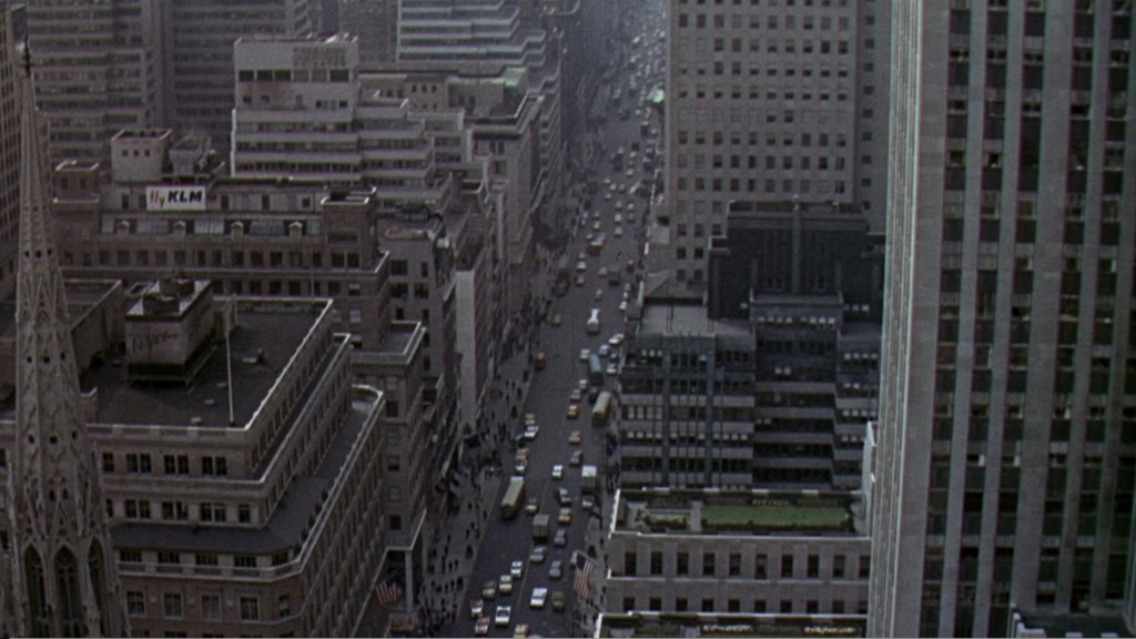 Aerial view of a busy city street with tall buildings, featuring a mix of modern and older architecture, and traffic moving along the road.
