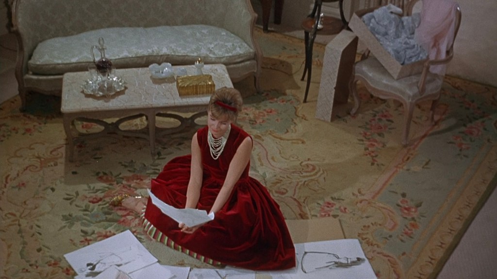 A woman in a red dress sits on the floor surrounded by sketches, looking at a piece of paper. A soft sofa and a small table with drinks are in the background, along with a box of clothing.