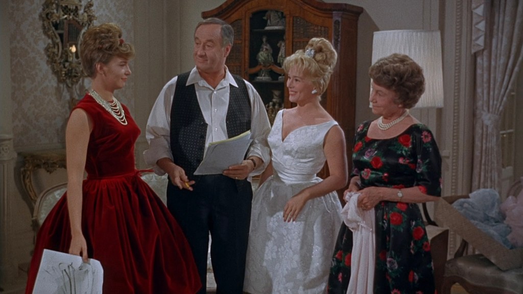 A scene featuring four people in a well-decorated room; two women in formal dresses, one in a red dress and the other in a white dress, stand beside a man holding a script, while another woman in a floral dress observes.