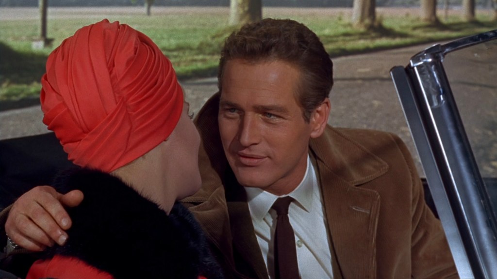 A man seated in a car looks intently at a woman beside him, who is wearing a bright red turban and black fur coat. The scene is set outdoors with trees visible in the background.