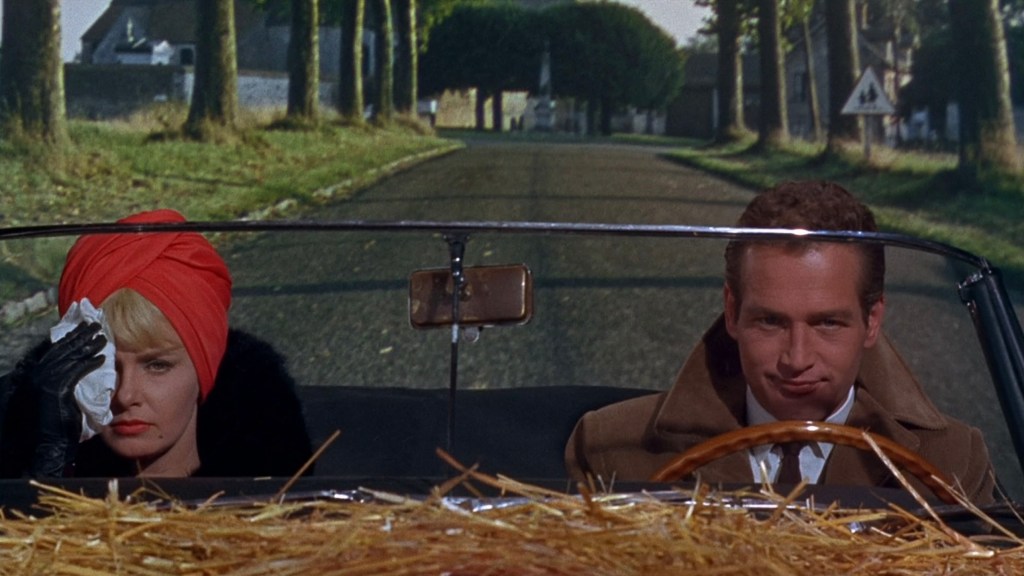 A man and a woman sitting in a convertible car on a rural road. The woman is wearing a red headscarf and black gloves, wiping her face with a tissue, while the man is smirking.