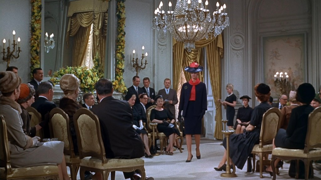 A fashion show scene featuring a model walking in a stylish outfit and hat, with an audience of elegantly dressed spectators seated around her in a lavishly decorated room.