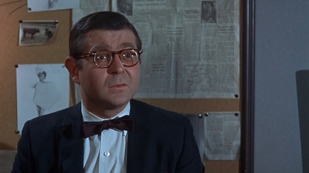 A concerned man in glasses and a bow tie, sitting in front of a corkboard filled with various papers and clippings.