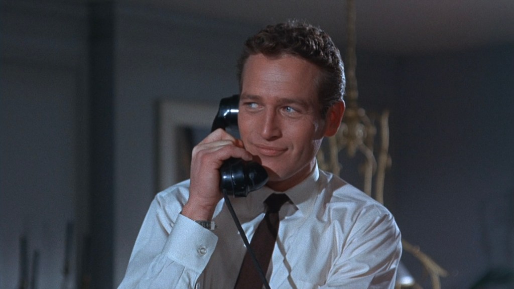 Man in a white shirt and tie speaking on a vintage black telephone with a slight smile.