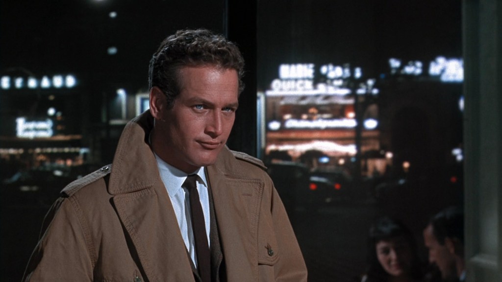 A man in a beige trench coat stands with a confident expression against a nighttime urban backdrop with blurred city lights.