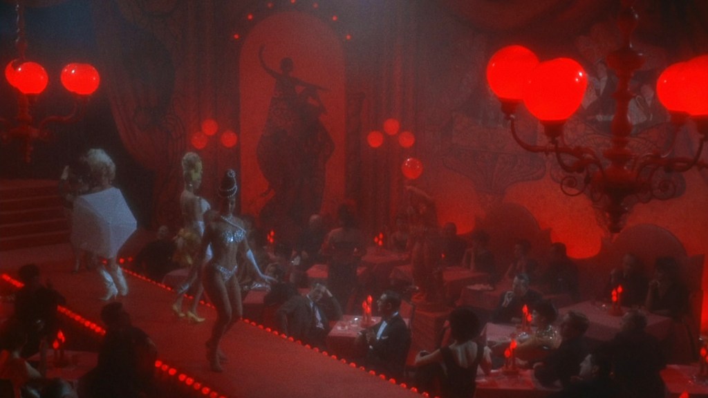 A vibrant nightclub stage with performers in extravagant costumes, illuminated by red lighting, while an audience watches at tables below.