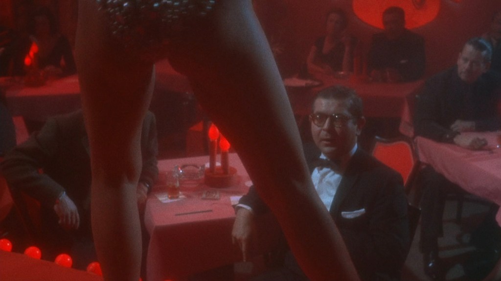 A dimly lit cabaret scene featuring a performer on stage, with a focus on their legs, while captivated audience members watch at tables adorned with red lighting.