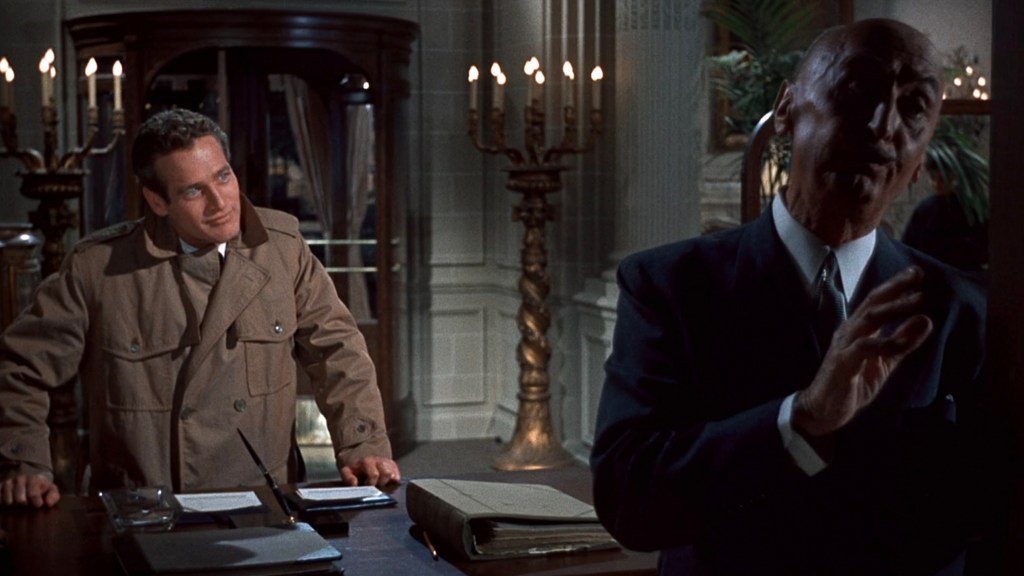 A scene featuring two men in a dimly lit room. One man, dressed in a beige coat, leans over a desk with papers, while the other man in a dark suit gestures as he speaks, surrounded by candelabras and ornate furnishings.