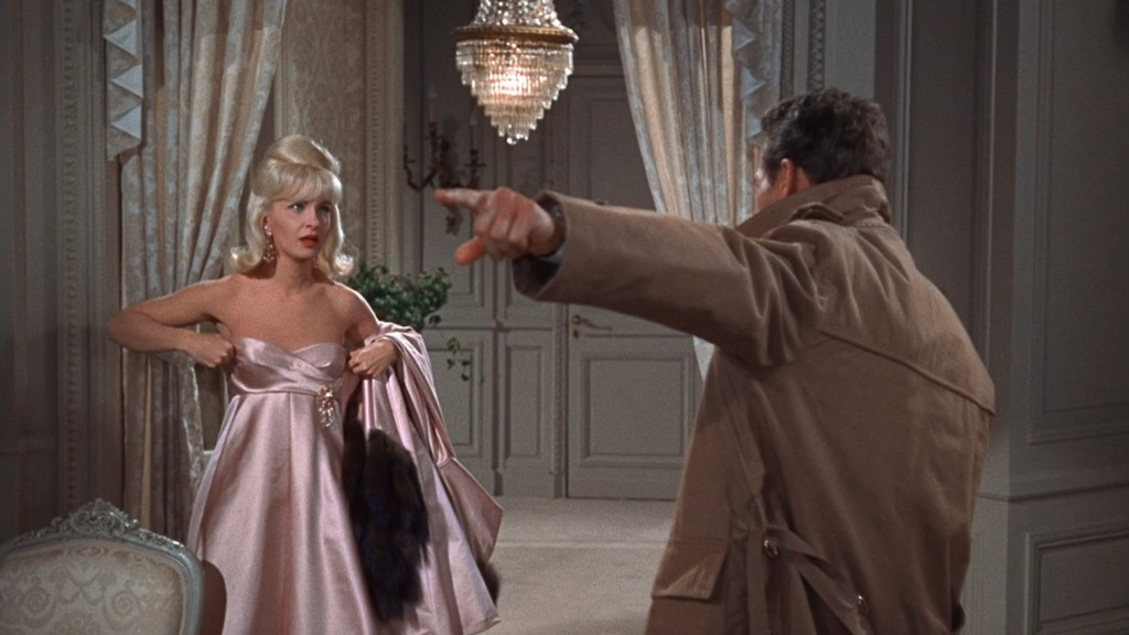 A blonde woman in a pink dress looks surprised while a man in a trench coat points at her, set in an elegantly decorated room with a chandelier.