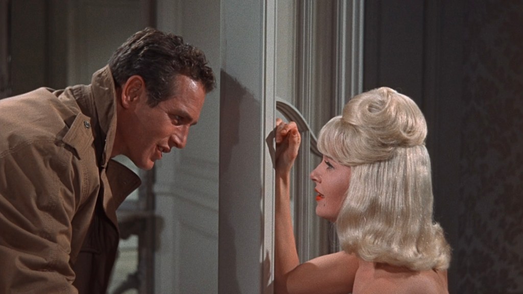 A close-up scene featuring a man in a coat leaning towards a woman with blonde hair, both appearing engaged in a conversation. The woman is resting her hand on a wall while gazing at the man.