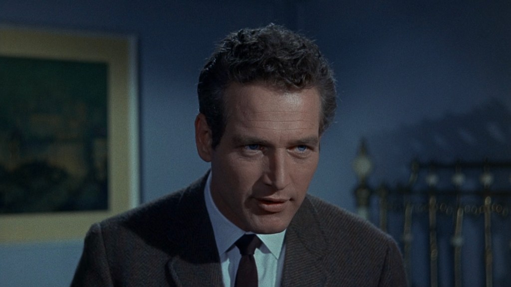 Close-up of a man with curly hair, wearing a suit and tie, engaged in conversation in a dimly lit room.