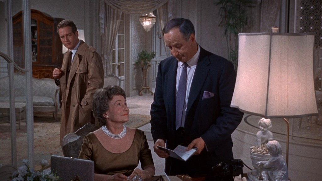 A woman sits at a desk in a stylish interior, smiling at a man in a suit who is standing nearby and reading a document. Another man in a trench coat appears in the background, observing the scene.