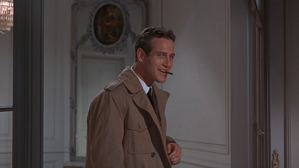 A smiling man in a beige coat, holding a cigar, stands in a well-decorated room with ornate details.