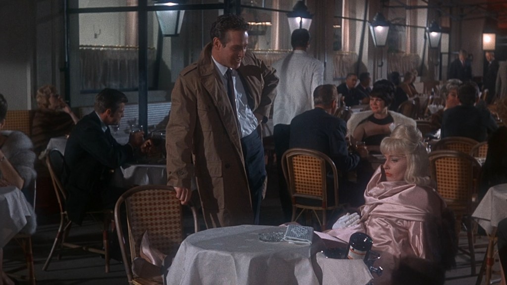 A man in a trench coat stands speaking to a woman in a pink shawl at a restaurant table, surrounded by other diners in an elegant setting.
