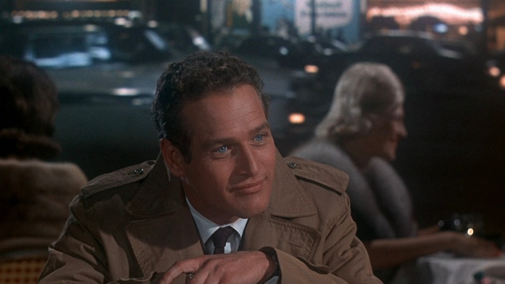 A man in a beige coat sitting at a cafe table with a slight smile, amidst a blurred lively city nightlife in the background.