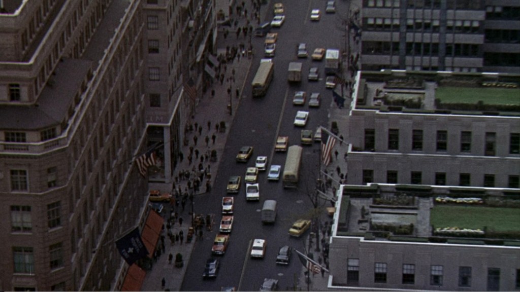 Aerial view of a busy city street filled with cars and pedestrians, featuring several buildings with flags and greenery in urban settings.
