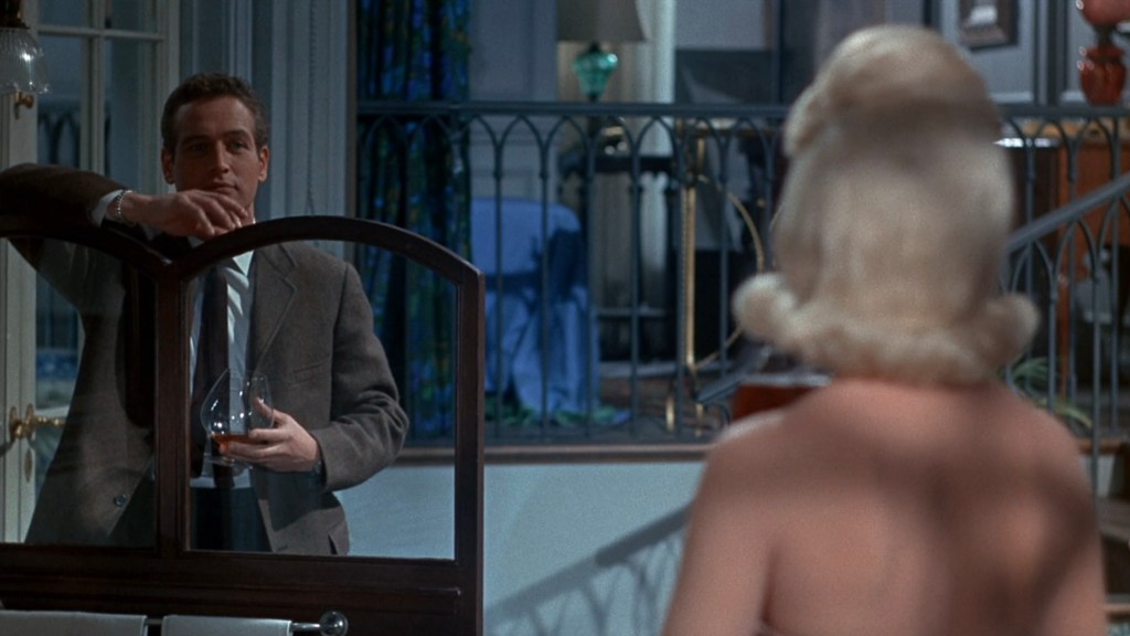 A man in a suit leans against a partition while holding a glass, looking at a woman with blonde hair, who is partially out of frame, in a well-furnished room with visible décor.