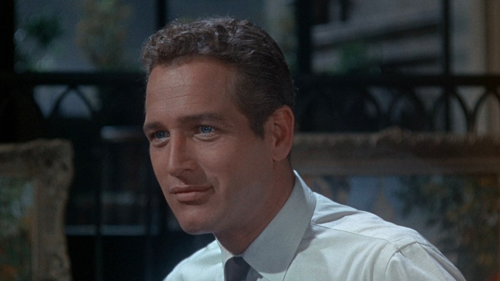 Close-up of a man with short, wavy hair, wearing a light-colored shirt and tie, smiling softly with a serious expression.