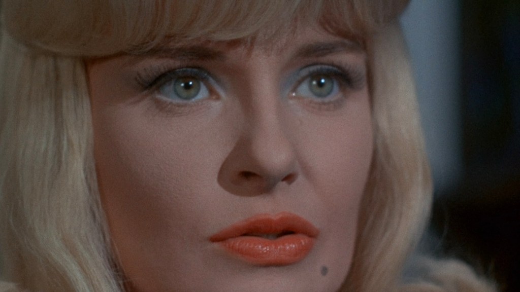 Close-up of a woman with blonde hair, blue eyes, and bright orange lipstick, expressing a serious or contemplative emotion.