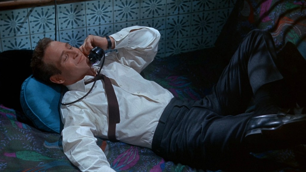A man in a white dress shirt and black pants relaxes on a patterned couch while talking on a vintage telephone.