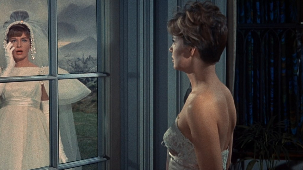 A bride in a white wedding dress with a veil stands outside a window, looking distressed while touching her face. Inside, a woman in a strapless dress gazes back at her.