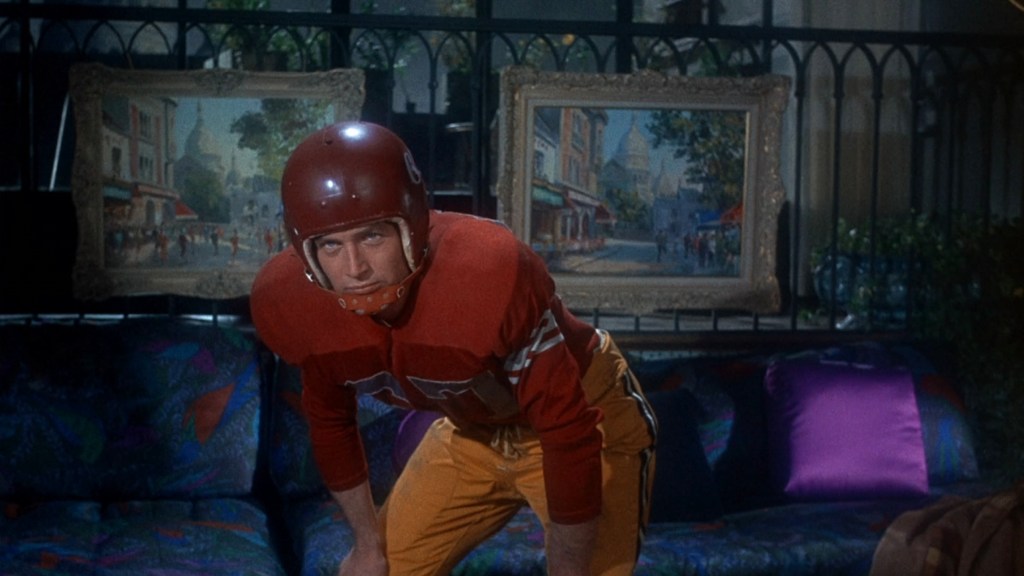 A man wearing a red football helmet and uniform is crouched on a colorful couch, with framed paintings visible in the background.
