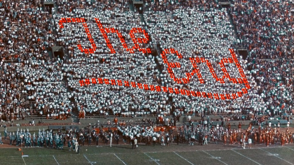 A large crowd at a stadium forming the text 'The End' using white and orange materials, with a marching band and performers in the foreground.
