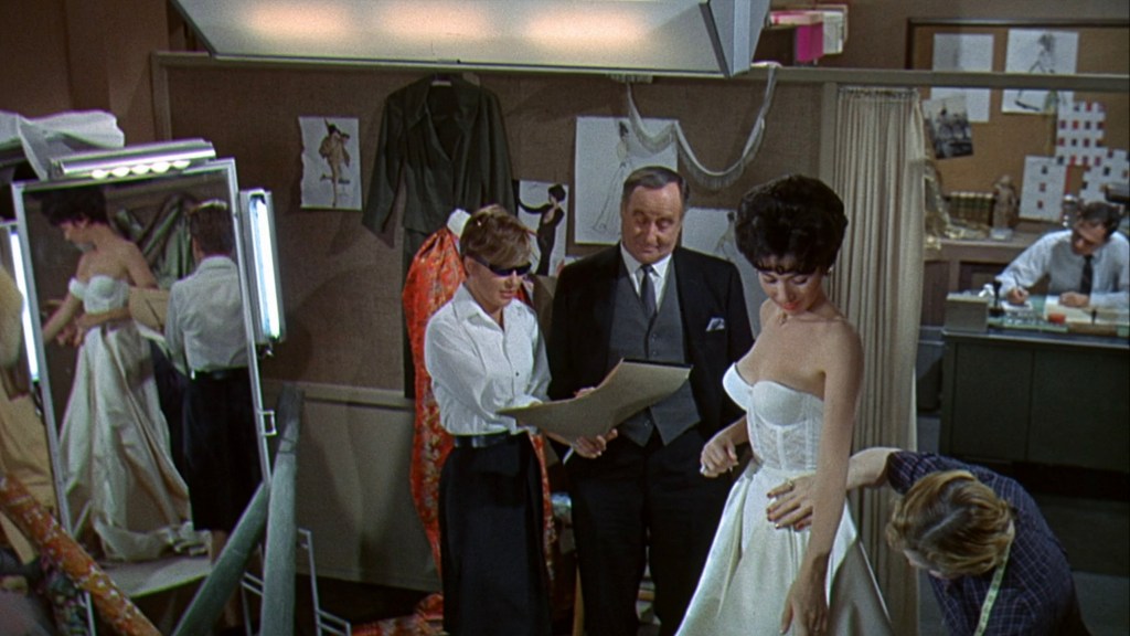 A fashion fitting scene featuring a woman in a white gown, while a designer and assistants provide feedback. A mirror reflects the scene, with sketches and garments visible in the background.