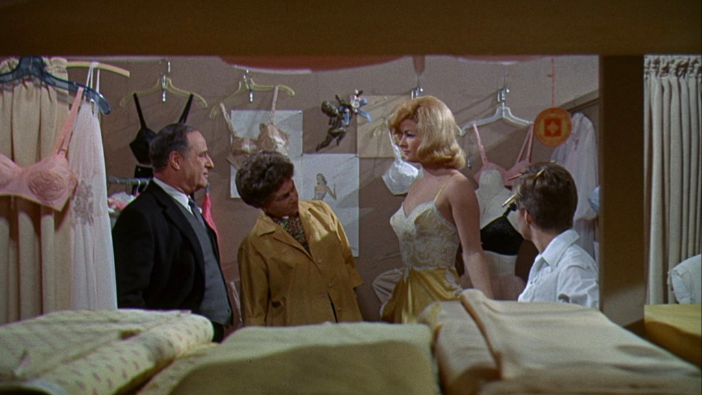 A scene in a clothing store featuring a group of four individuals, including a woman trying on a dress, while others observe and discuss. The background is filled with various garments displayed on hangers.