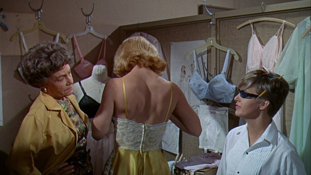 A scene in a clothing store featuring three women. One woman in a yellow dress is trying on clothing while another woman observes her from the side. The third woman, wearing glasses, is taking notes or measuring.