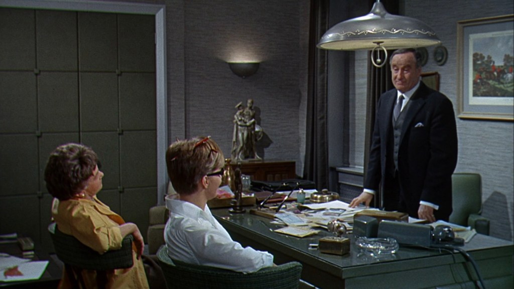A scene in an office featuring three characters: a man in a suit standing at a desk, and two individuals sitting across from him, one wearing glasses and the other in an orange outfit. The room is furnished with a large desk, an overhead light, and a decorative statue in the background.