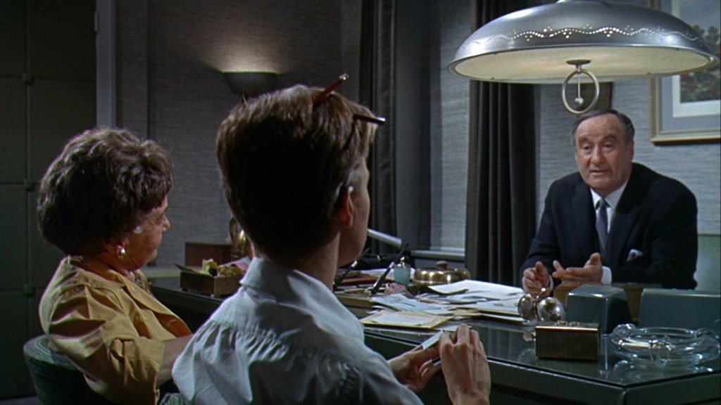 A scene in an office with three people: two sitting at a desk, one facing the camera and the other slightly turned, while a man in a suit speaks from the other side of the desk under a pendant lamp.