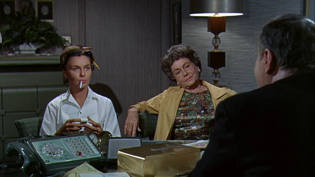 A woman with a cigarette sits casually at a table, looking contemplative, while another woman in a colorful outfit leans back, observing a man speaking across from them in an office setting.