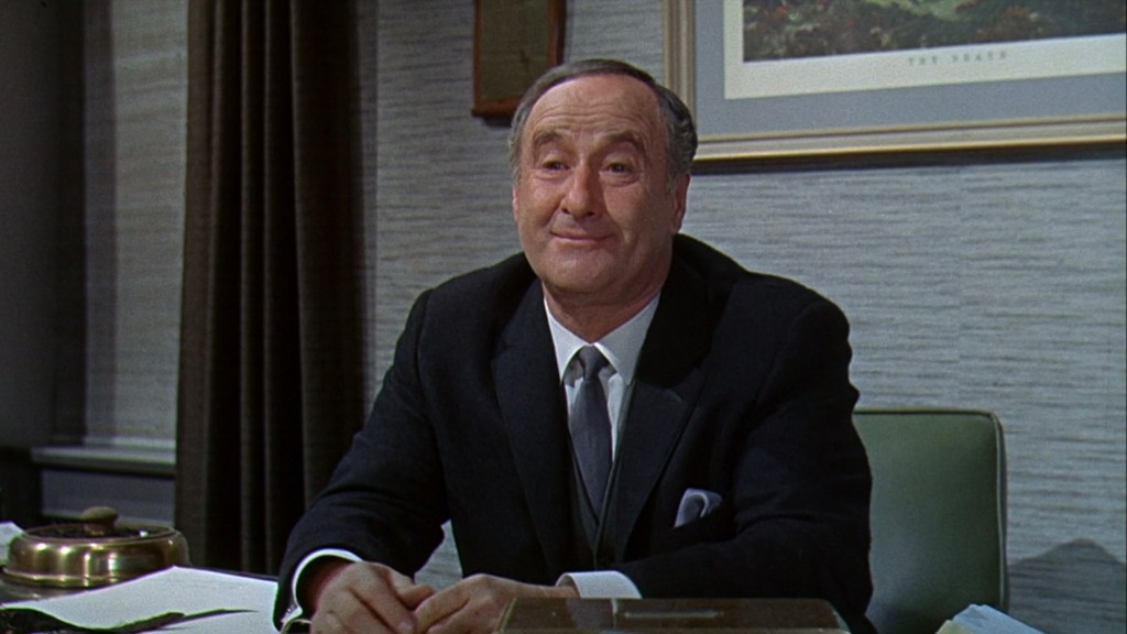 A man in a suit smiling while sitting at a desk, with a framed picture on the wall and curtains in the background.