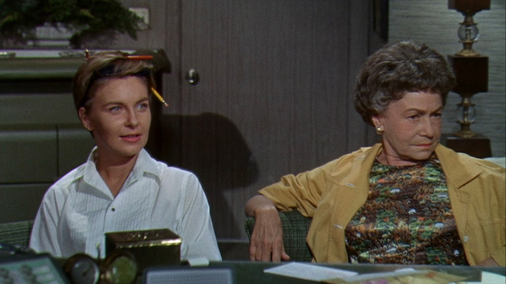 Two women sitting at a table, one with a pencil in her hair and wearing a white shirt, the other with short curly hair in a yellow jacket and patterned top.