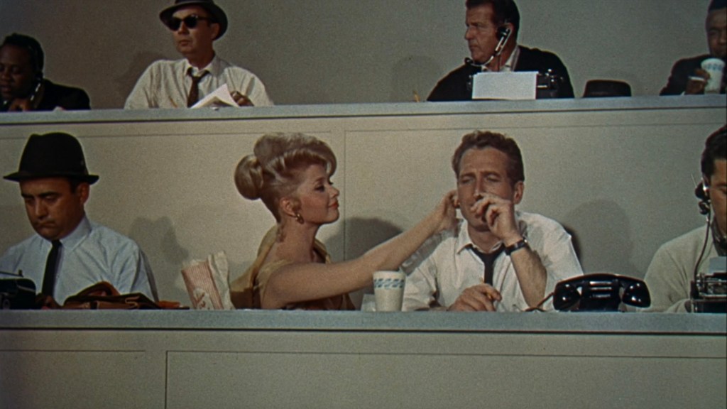 A woman gently touches a man's face while he smokes a cigarette in a crowded press box, with other reporters seated behind them.