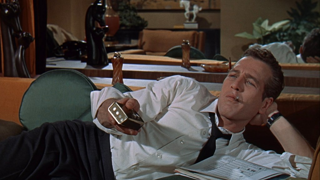 A man lounging on a couch with a remote control, wearing a white shirt and black tie, looking thoughtful while smoking a cigarette.