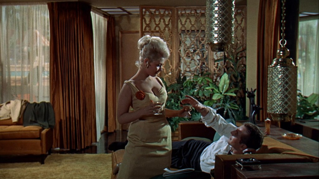 A stylish woman in a yellow dress holding a drink stands beside a man lounging on a couch in a modern interior setting with plants and decorative lighting.